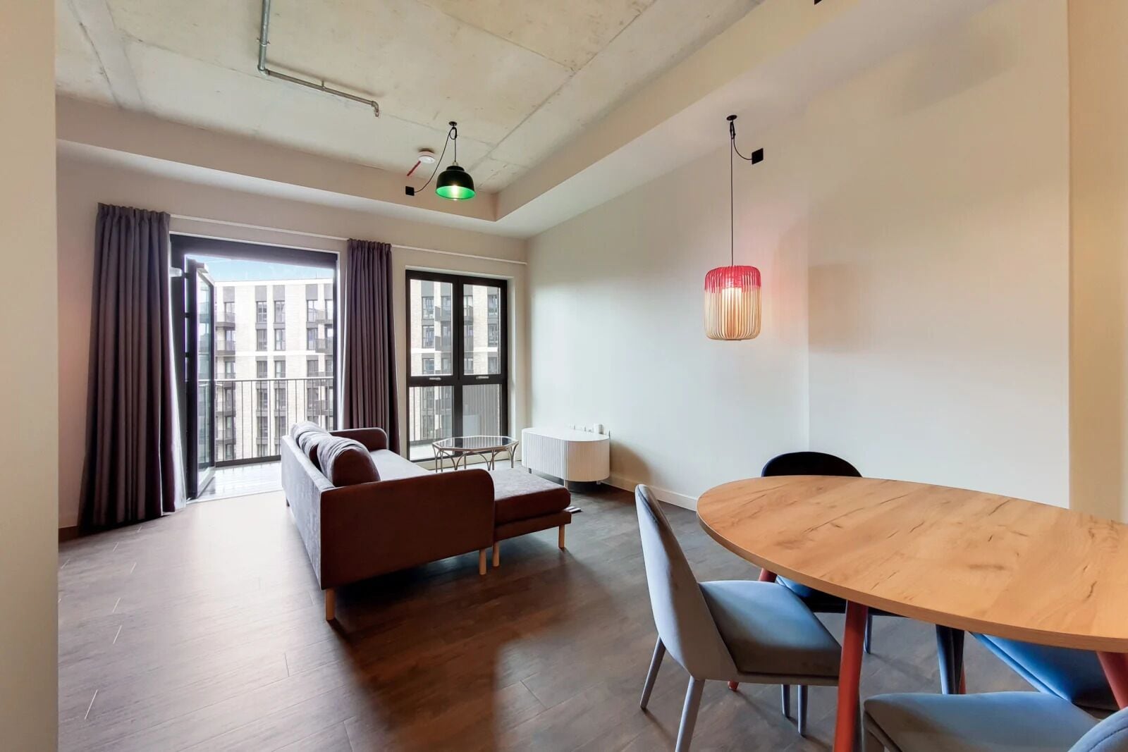 Een moderne kamer met een houten vloer, een bruine bank, een ronde houten tafel en een groot raam met gordijnen.