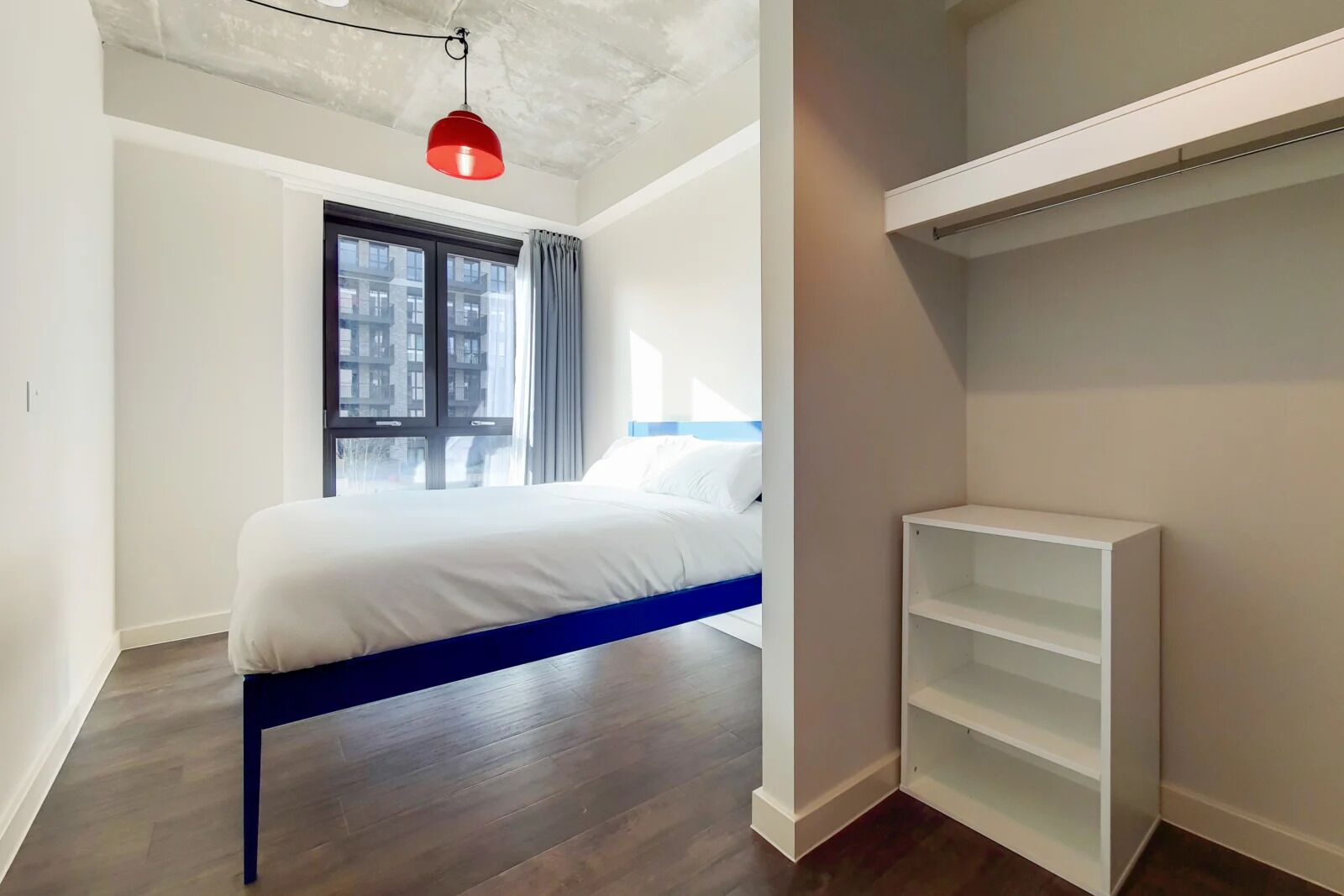 Een slaapkamer met een bed, een raam, een rode hanglamp en een witte wandplank.
