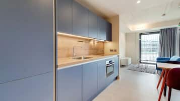 A modern kitchen with grey cabinets, a stainless steel sink, and a built-in oven. There is a dining area with a table and chairs, and a large window with curtains.