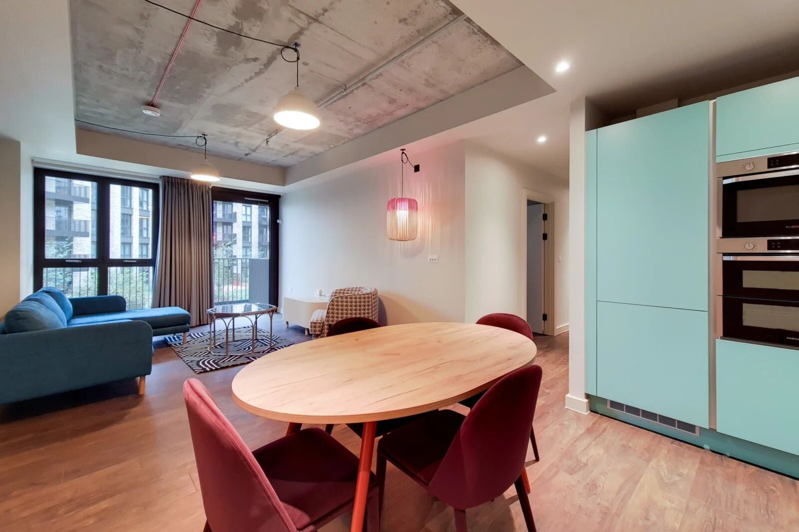A modern living room with a sofa, a round wooden table, and red chairs. There is a large window with curtains, a ceiling with exposed beams, and a kitchen area with cabinets.