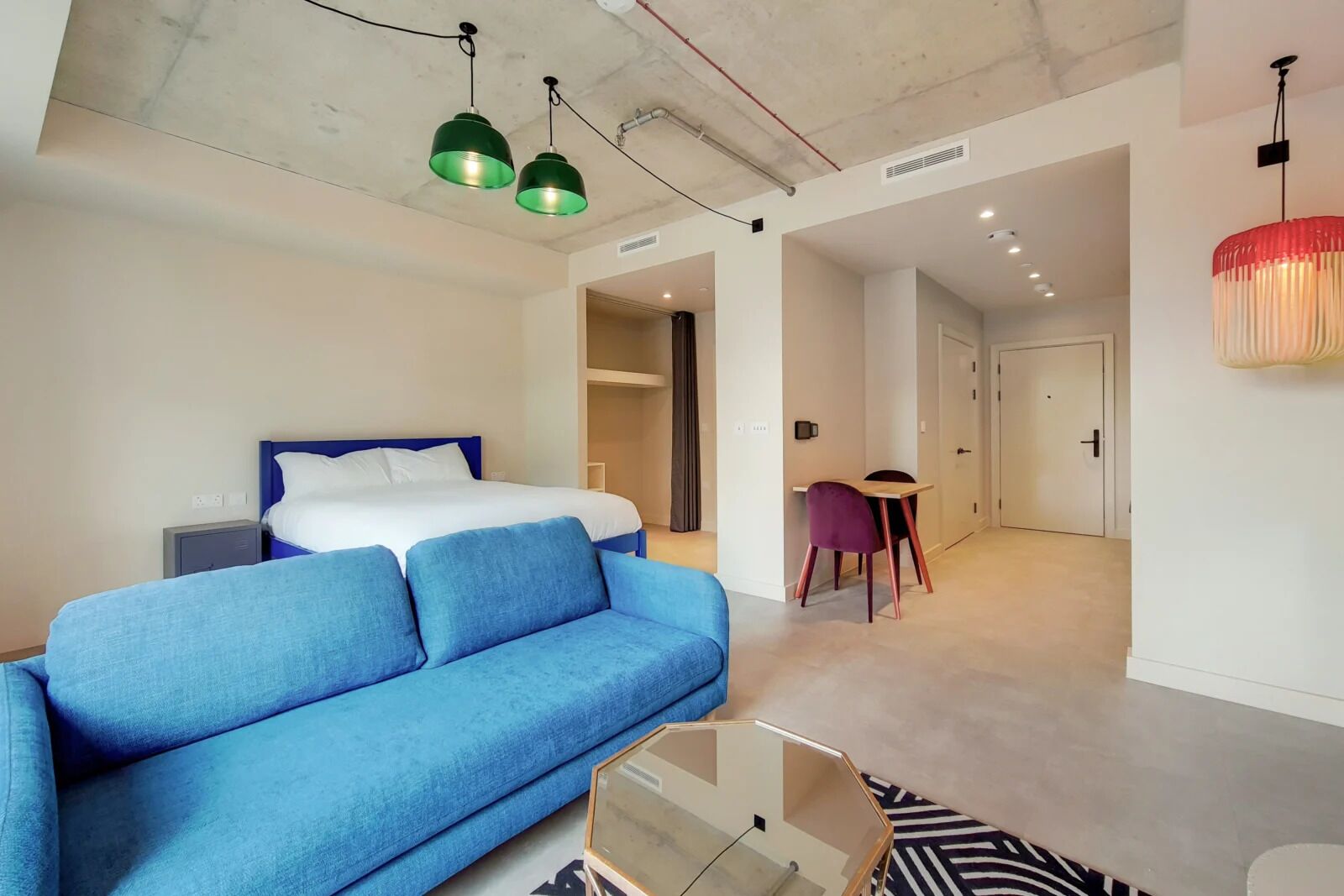 A modern hotel room with a bed, blue sofa, and a dining area with a table and chairs.