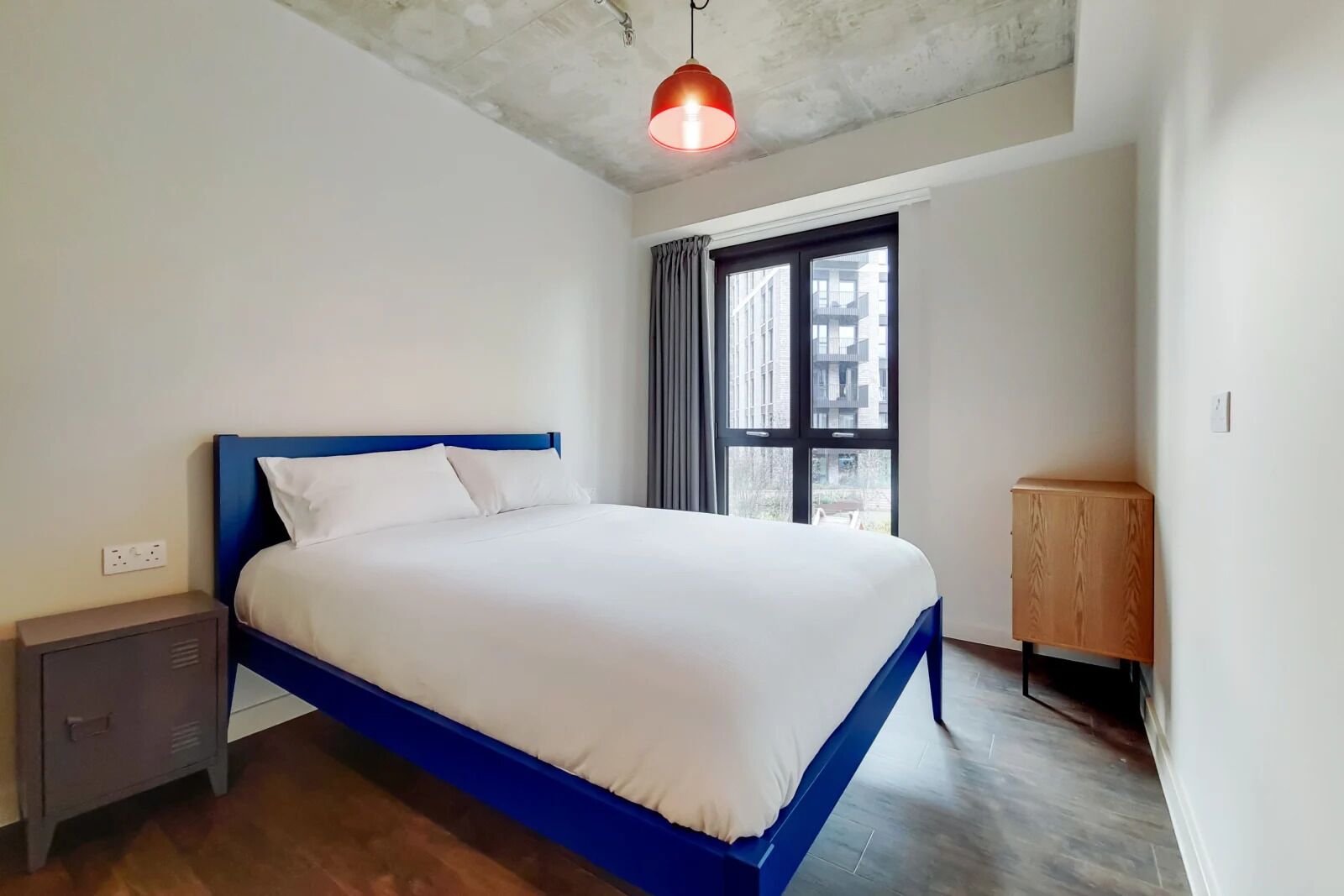 A bedroom with a bed, a nightstand, a wooden cabinet, a window with curtains, and a red pendant light.