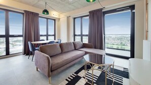 A modern living room with a sofa, a dining table with chairs, large windows with curtains, and a view of the city. - The Robinson (Wembley)