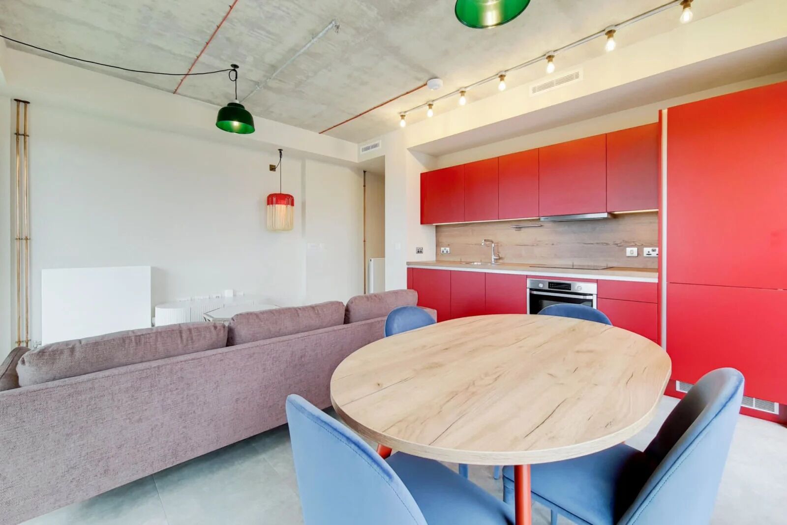 A modern living space with a round wooden table, blue chairs, a grey sofa, and red kitchen cabinets.