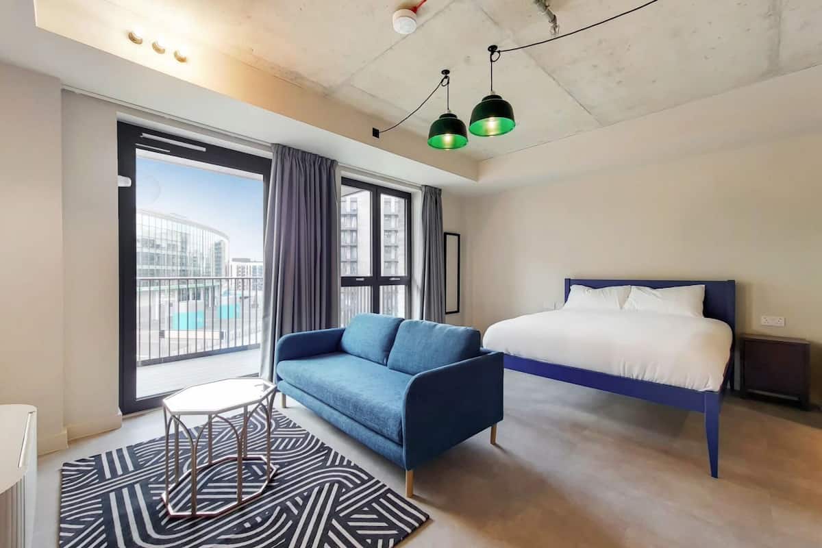 Quarto de hotel moderno com uma cama grande, um sofá azul, uma pequena mesa e vista para a cidade através de amplas janelas.