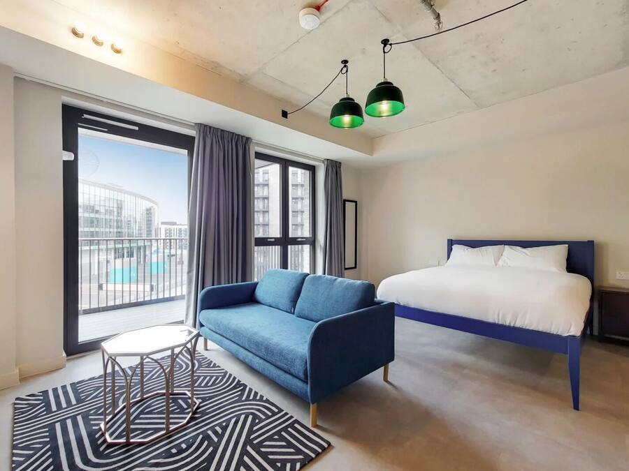 A modern hotel room with a large bed, a blue sofa, a small table, and a view of the city through large windows.