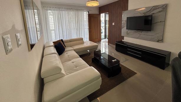 Un salon moderne avec un canapé d’angle, une table basse en verre, un téléviseur à écran plat fixé au mur et un grand miroir.