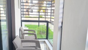Un balcon agrémenté de meubles en osier, d’un luminaire au design géométrique et offrant une vue sur un immeuble et la verdure.