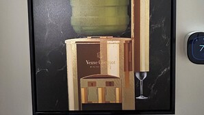 Ein gerahmtes Kunstwerk mit einer grünen Flasche, einer Holzkiste und einem Weinglas an einer Marmorwand.