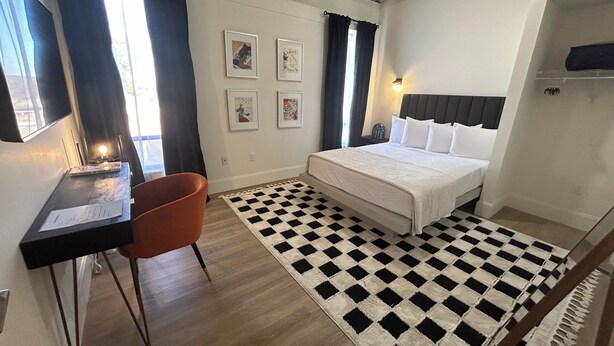 Ein Hotelzimmer mit Bett, Schreibtisch, Stuhl, Teppich mit Schachmuster und gerahmten Bildern an der Wand.