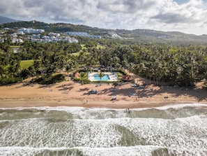Una playa con zona de piscina, palmeras y edificios al fondo.