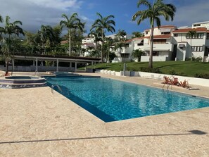 Ein Pool mit gefliestem Sonnendeck, umgeben von Palmen und einem überdachten Bereich mit Stühlen.