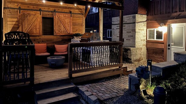 Terraza exterior con mobiliario de madera, una parrilla y luces colgantes.