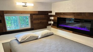 Un dormitorio con cama, dos almohadas, una ventana con vista a follaje y un televisor montado en la pared.