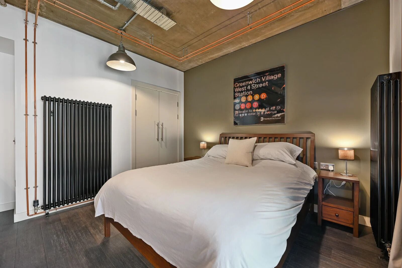 A bedroom with a large bed, wooden headboard, two bedside tables with lamps, a radiator, and a poster on the wall.