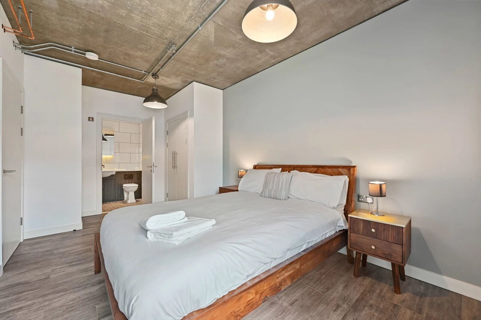 Une chambre à coucher moderne avec un lit en bois, du linge de lit blanc, une table de chevet avec une lampe, et une salle de bain visible à travers une porte ouverte.