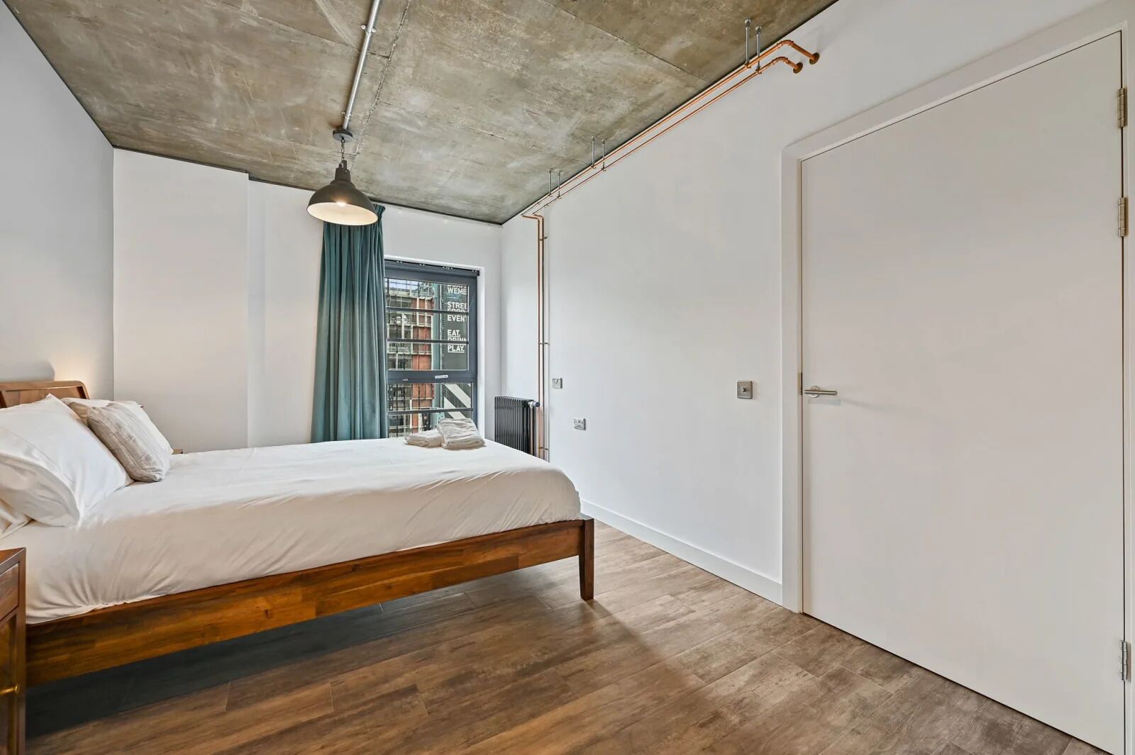 A bedroom with a wooden bed, a hanging light fixture, a view of the city through a window, and a white door.