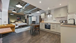 A modern kitchen with white cabinets, a built-in oven, and a sink. There is a dining area with a table and chairs, and a bed in the adjacent room.