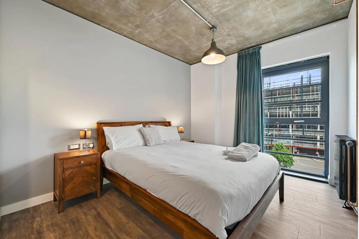 Ein modernes Schlafzimmer mit einem hölzernen Bett, Nachttischlampen, Blick auf eine Baustelle und einer Heizung.