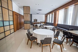 A modern dining area with round tables and chairs, a long bench, and a kitchen in the background. - Grand Flora Hotel (Melaka)