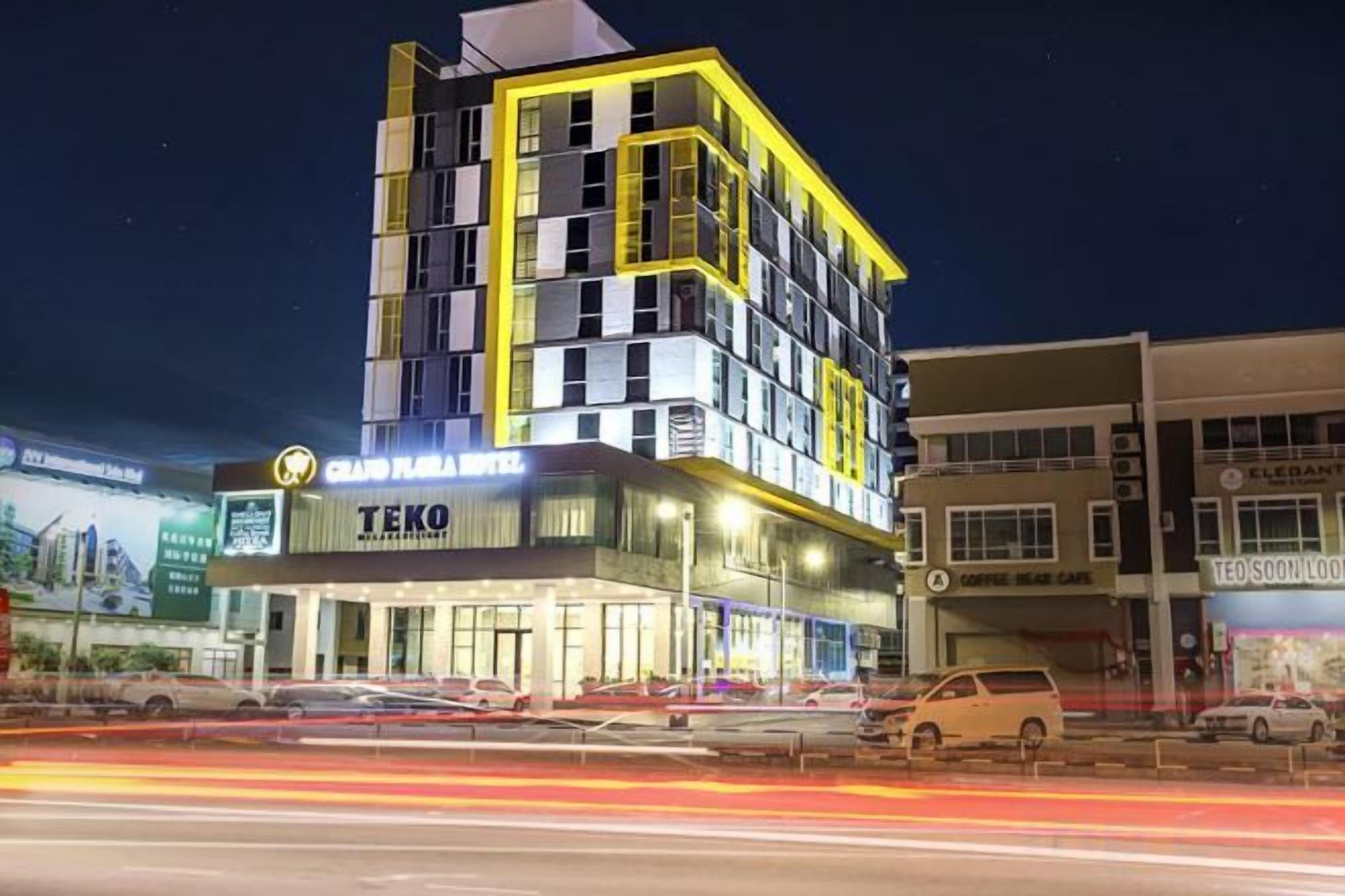 A multi-story hotel with a prominent 'TEKO' sign, a parking lot with cars, and a nearby building with a 'TEOS' sign.