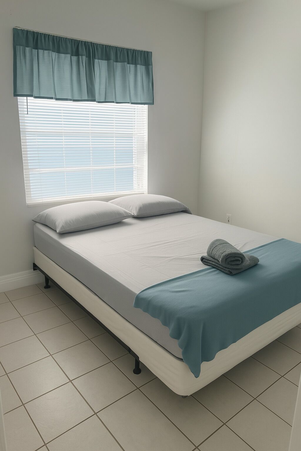 A bedroom with a bed, a window with blinds, a teal valance, and a rolled towel on the bed.
