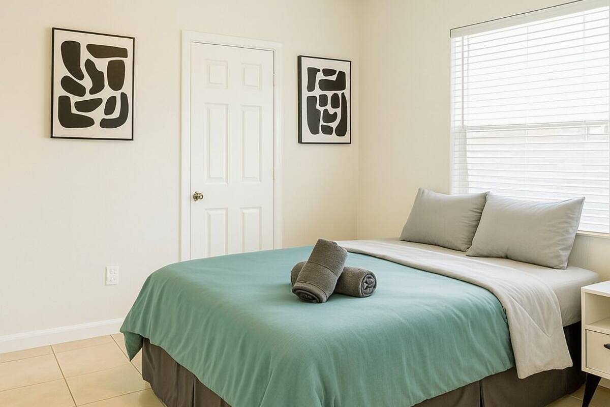 A bedroom with a bed, a nightstand, a window with blinds, and two abstract art pieces on the wall.