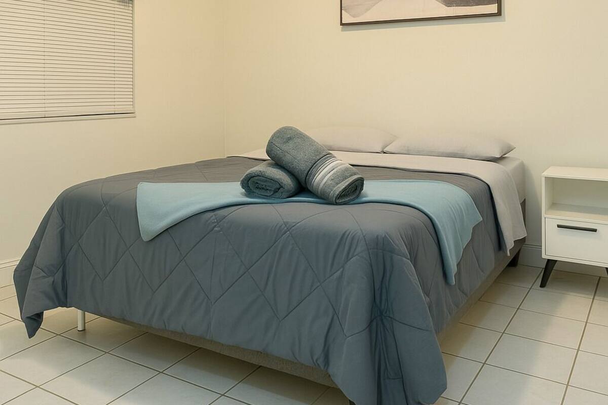 A bedroom with a bed, a nightstand, a wall-mounted light, a window with blinds, and a painting on the wall.