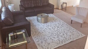 A living room with a dark brown leather sofa, a glass coffee table, a patterned rug, a round metal table, a ceiling fan, a mirror, and a white chair. - Charming 2-bedroom condo with WiFi, AC in delightful Canton (Canton)