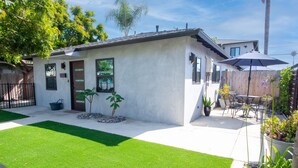 A modern house with a white exterior, a small front yard with artificial grass, and a patio area with outdoor furniture and a large umbrella.