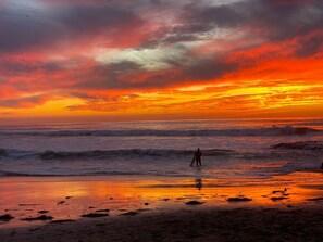 Escena de playa con una persona de pie en el agua, olas y un atardecer.