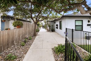A narrow concrete pathway flanked by a wooden fence and a black metal fence, leading to a white building with a dark roof.