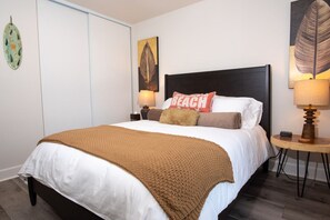 A neatly made bed with a brown blanket, white pillows, and a decorative pillow with the word "BEACH". There are two wall-mounted lamps on either side of the bed.