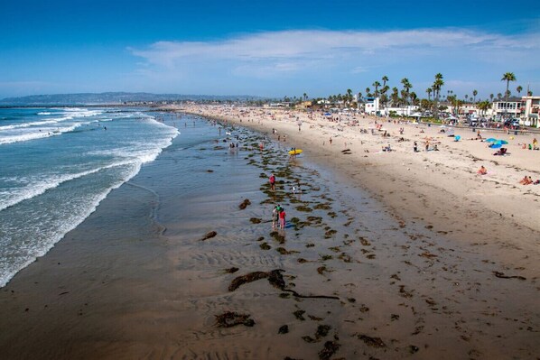 A wide beach with clear blue water, palm trees, and buildings in the background.