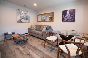 A modern living room with a grey sofa, a round coffee table, and a glass-top dining table with wooden chairs. There are abstract paintings on the walls and a mirror above the sofa.