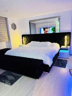 Un dormitorio moderno con una cama negra, ropa de cama blanca, un espejo grande sobre la cama y una mesita de noche con una lámpara.