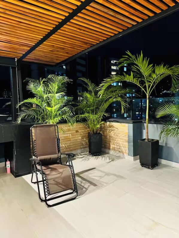 Una terraza en la azotea con techo de madera, plantas en macetas y una silla plegable.