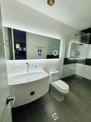 Un baño moderno con un espejo grande, un lavamanos blanco, un inodoro y una zona de ducha con mampara de vidrio.