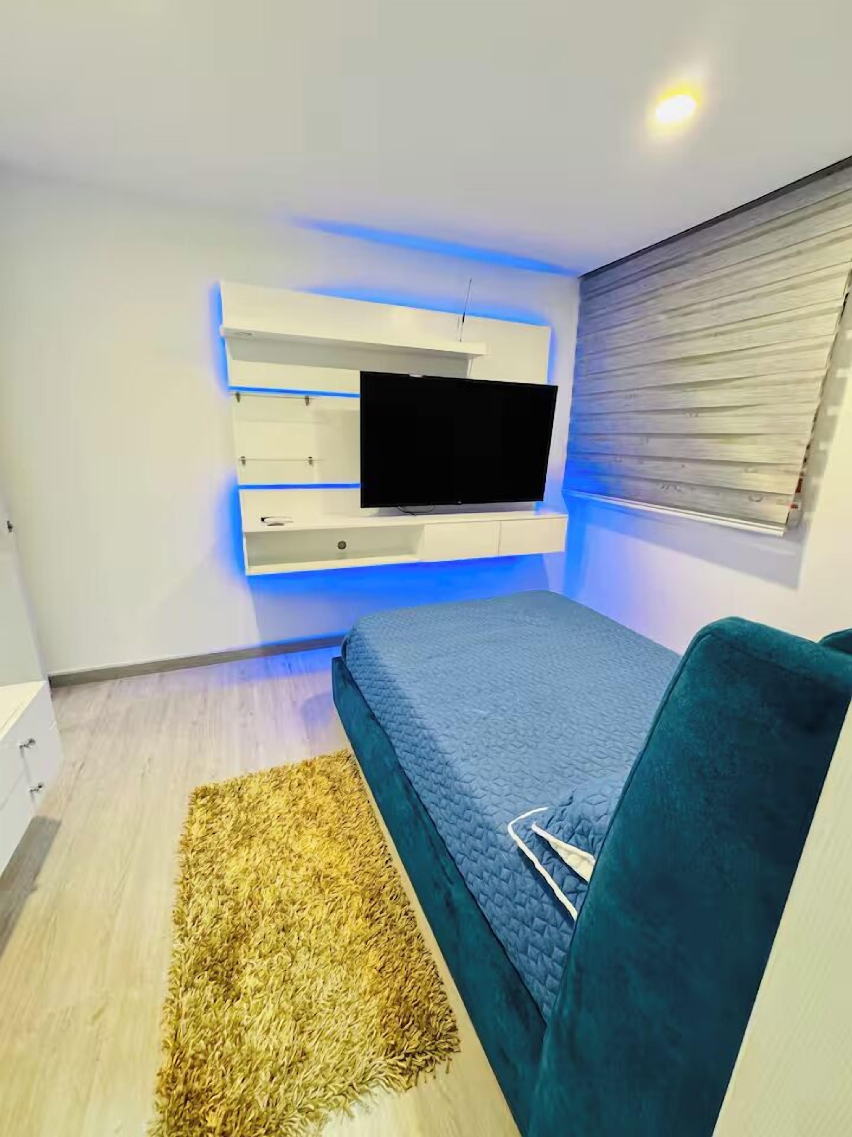 Un dormitorio con un televisor montado en una pared blanca, una colcha azul y una alfombra marrón en el suelo.