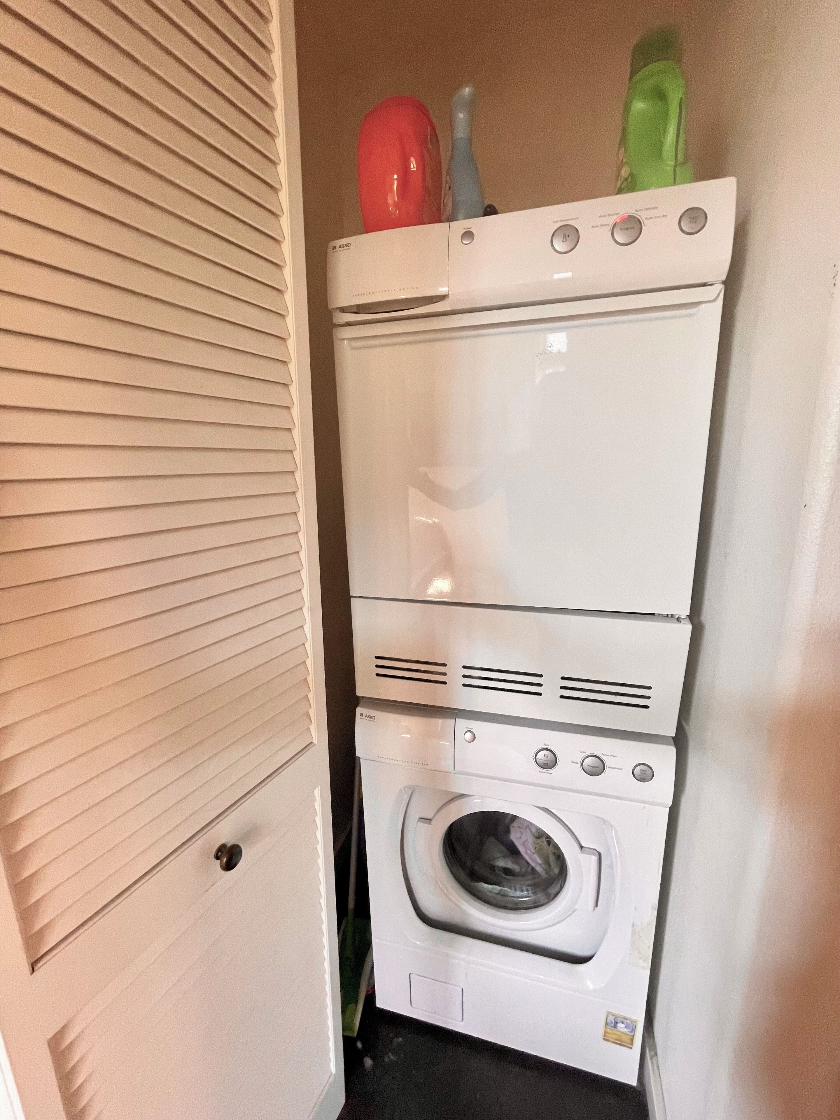 Ein kleiner Hauswirtschaftsraum mit einer gestapelten Waschmaschine und einem Trockner, einer Tür mit Griff und einem an der Wand befestigten Regal mit einem roten und einem grünen Gegenstand.