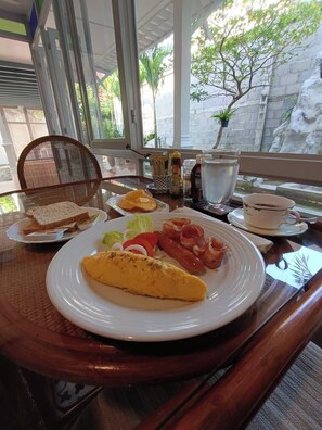 A breakfast setting with a plate of eggs, bacon, and toast, a cup of coffee, and a glass of water on a table with a wicker chair and a view of a stone wall and greenery outside. - Baan Debtida (Bangkok)