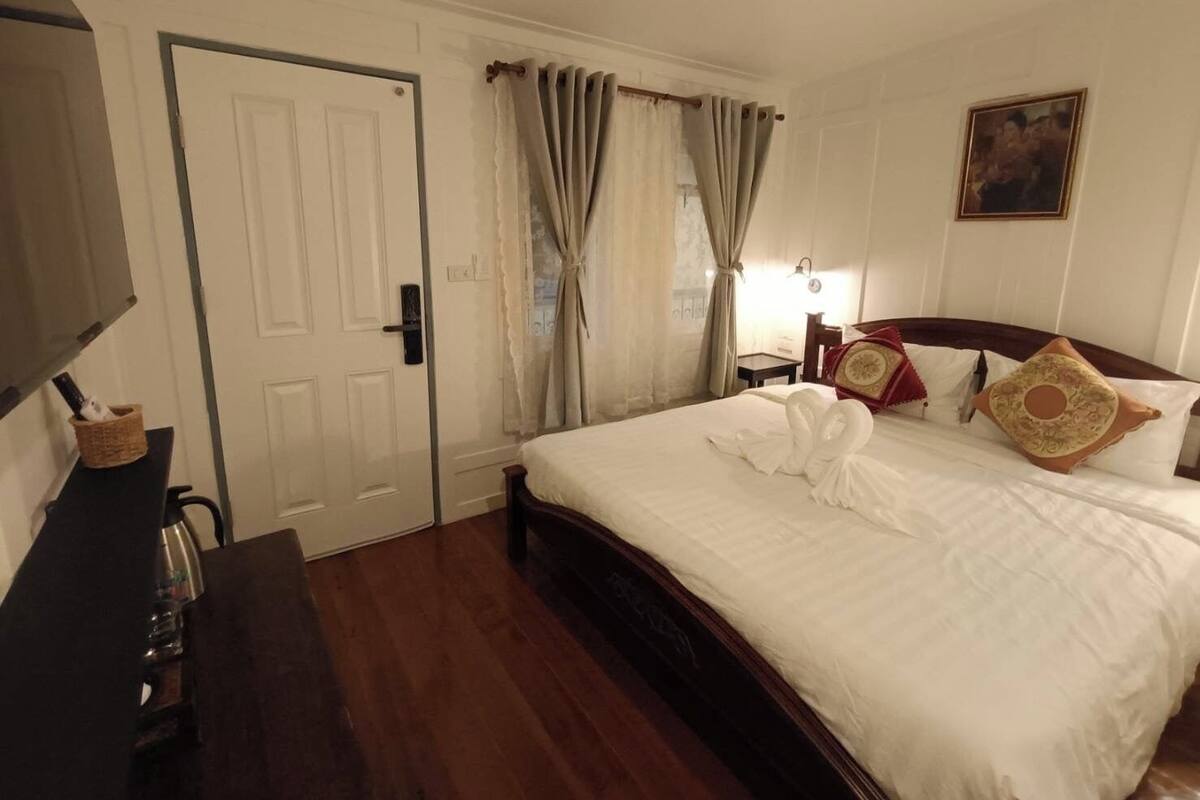 Une chambre d’hôtel avec un lit en bois, des rideaux blancs, un téléviseur à écran plat et une petite table de chevet.