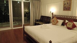Ένα δωμάτιο ξενοδοχείου με ένα μεγάλο κρεβάτι, ένα ξύλινο προσκέφαλο, ένα κομοδίνο και μια καρέκλα. Υπάρχει μια συρόμενη γυάλινη πόρτα που οδηγεί σε έναν εξωτερικό χώρο.
