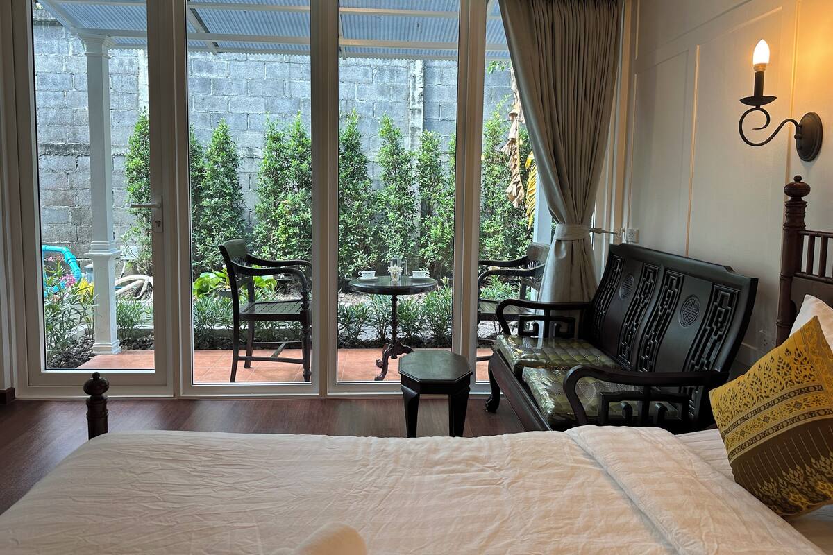 Une chambre d’hôtel avec un grand lit, un banc en bois, une petite table et une vue sur un jardin, visible à travers des portes vitrées.