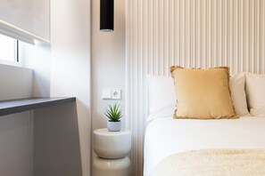 A modern hotel room with a bed, a bedside table with a plant, a wall-mounted light, and a window. - Calma Rooms by Charming (Madrid)