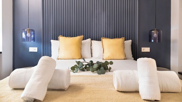 A neatly made bed with white linens and yellow pillows, a beige rug, and a plant on the bed. - Calma Rooms by Charming (Madrid)