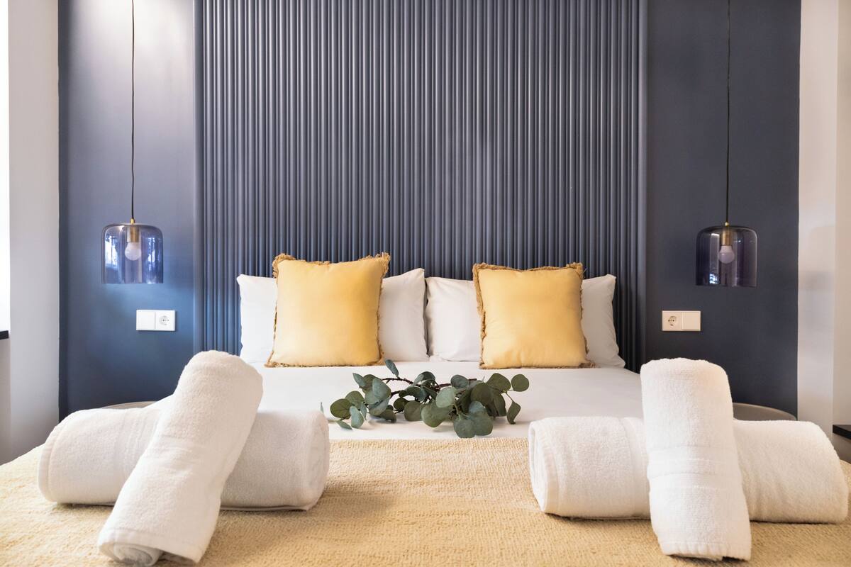 A neatly made bed with white linens and yellow pillows, a beige rug, and a plant on the bed.