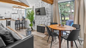 A modern kitchen with white cabinets, a dining area with a round wooden table, and a TV mounted on a textured stone wall.