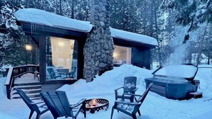 A cabin with a stone chimney, outdoor seating, and a hot tub in a snowy setting.