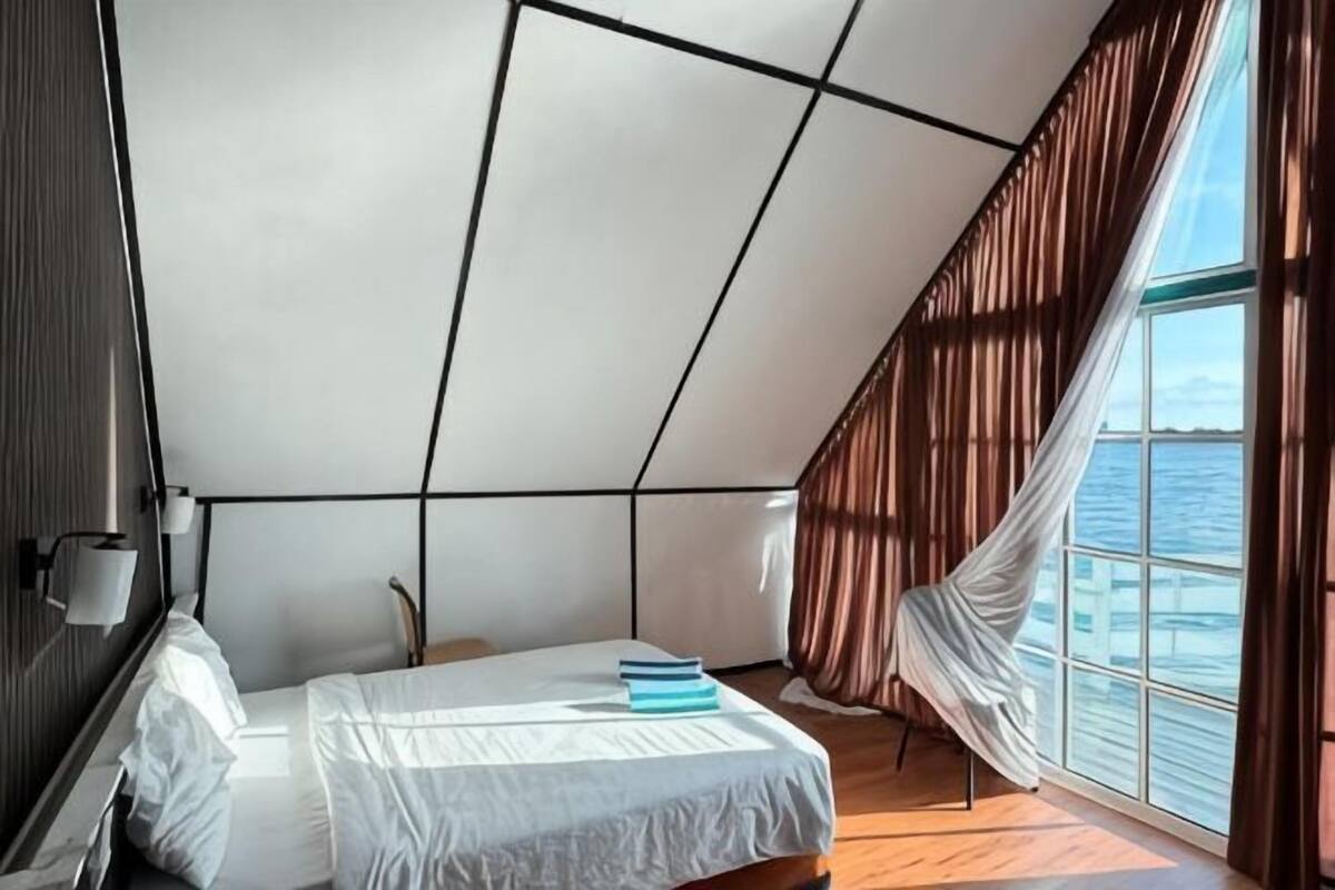 A bedroom with a bed, a window with curtains, and a view of the sea.